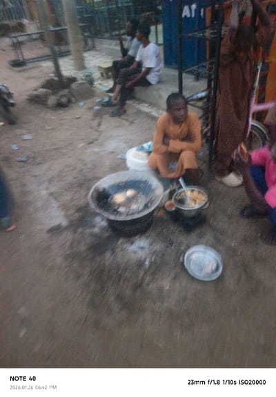 This image captures an outdoor, evening scene in Panshekara, Nigeria, depicting what appears to be a street food operation. In the foreground and center, a person dressed in an orange garment is seated on the ground, attending to food cooking in a large, dark pan or wok placed over a makeshift stove on the dirt ground. Two round, white objects, possibly eggs or dough balls, are visible in the pan. Next to the large pan, a smaller pot containing light-colored food, possibly fried items, is also present. An empty, clear plastic plate lies on the ground to the right.
Several other individuals are present. In the mid-ground, two men are seated on a raised concrete platform or curb. One wears a white t-shirt and dark trousers, while the other, further back, wears a lighter shirt. A blue metal stand or cart with the letters "AT" visible on it stands behind them. On the far right, a person in a brown garment stands or sits beside the blue cart, and another person wearing a pink shirt is partially visible, holding an object. The ground is unpaved, covered in dirt, and scattered with what appears to be litter or small pieces of paper.
The overall lighting is dim, consistent with the timestamp of "06:42 PM" on January 26, 2026. The entire image is quite blurry, likely due to a slow shutter speed (1/10s) and high ISO (ISO20000) used in low light conditions. The bottom of the image displays camera metadata: "NOTE 40", "2026.01.26 06:42 PM", and "23mm f/1.8 1/10s ISO20000".