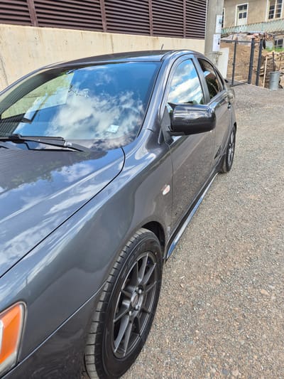 An exterior, eye-level, close-up shot captures the driver's side of a dark gray sedan parked on a gravel surface. The car is angled slightly towards the viewer's right. The left side of the vehicle is in focus, revealing the front fender, wheel, door, and side mirror. The car's paintwork is highly reflective, mirroring the blue sky and white clouds above. The side mirror features a carbon fiber casing, a visual contrast to the car's body.
In the background, a building with a light-colored facade and multiple windows is visible. A fence made of vertical wooden planks stands in front of the building. To the left of the car, a beige concrete wall with horizontal brown slats runs along the top edge of the frame. The environment appears to be a rural or semi-urban area, consistent with the town of Iskrets, Bulgaria, which is characterized by such architecture and unpaved surfaces. The lighting suggests it is daytime, with clear skies. No people are present in the image. The overall scene is static, with no apparent action taking place.