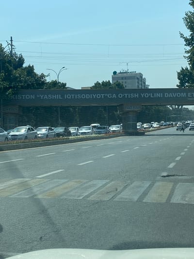 The image shows a street scene in Andijan City, Uzbekistan, on a clear, sunny day. A pedestrian bridge spans the road, with a banner displaying text in Uzbek. The banner reads "XISTON "YASHIL IQTISODIYOT"GA O'TISH YO'LINI DAVOM ET" which translates to "Continuing the path of the Green Economy". Below the banner, on the bridge supports, are decorative patterns.
The road is a multi-lane highway with moderate traffic. Several cars are visible, mostly white sedans, stopped or moving slowly. A pedestrian crossing is marked on the road in the foreground, with yellow and white stripes. On the right side of the road, trees line the median. In the background, a tall building with the logo "UZBEKINVEST" is visible, along with utility poles and overhead wires. The sky is a clear blue. There are no explicit interactions or specific actions of people clearly discernible other than the presence of drivers in the cars and possibly pedestrians further down the road. The overall impression is a typical urban road environment with a prominent infrastructural element and advertising.