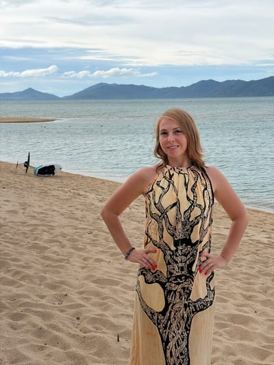 A medium-shot, eye-level, full body portrait of a Caucasian woman standing on a sandy beach in Ko Samui District, Thailand. The woman is smiling and looking at the camera. She has shoulder-length blonde hair and is wearing a long, sleeveless, light yellow dress with a black tree-like pattern. Her hands are on her hips. Behind her, to the left, is a small boat with a propeller on the sand near the water. The ocean is visible with mountains in the background, and the sky is partly cloudy.