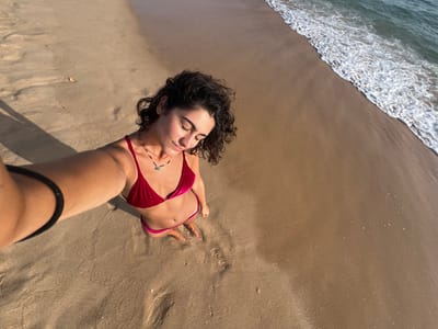 The image is a high-angle, vertical selfie of a female individual on a sandy beach near Almancil, Portugal. The subject, possessing dark, curly hair, is centrally positioned, with her head tilted downward and eyes closed. She is attired in a deep red velvet two-piece bikini and a necklace featuring a small, dark pendant. Her right arm is extended, holding the camera or phone for the selfie, with a dark band visible on her right wrist. Her bare feet are visible, standing on the light brown sand. To the right, the ocean is visible, characterized by green-blue water and white, foamy waves breaking onto the shore. A distinct, elongated shadow cast by the individual's arm and body extends diagonally across the sand from the lower left, indicating a low sun angle.