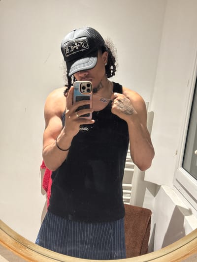 A person is reflected in an oval mirror, taking a selfie with an iPhone. The person is wearing a black trucker hat with silver embellishments, a black tank top, and navy striped pants. They have a silver necklace and a silver ring on their finger, and a black bracelet on their wrist. There are visible tattoos on the person's hand and arm. The mirror reflects a white wall, a brown towel, and part of a window with white trim. The location, Massy, France, is not discernible from the image alone.