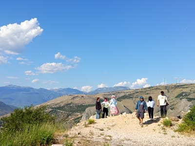 This photograph captures a group of six individuals on a hike amidst a mountainous landscape. The scene depicts a clear, bright blue sky with scattered white clouds, suggesting a pleasant, sunny day, likely in the late morning or early afternoon given the lighting.
The group consists of three women and three men, all dressed casually for outdoor activity. Some are walking forward, while others appear to be conversing or taking in the view. One woman in the center wears a distinctive pink and white floral skirt and a light blue headscarf, adding a touch of vibrancy to the group. Another woman is wearing a headscarf and a light blue jacket. The men are wearing t-shirts, shorts, and one is in a denim jacket. They are walking on a rocky, uneven path that overlooks a vast valley.
The background features rolling hills covered in dry grass and sparse vegetation, with layers of mountains stretching into the distance. The rugged terrain suggests a somewhat remote or wild natural environment. In the far distance, three wind turbines are visible on a hilltop, indicating the presence of human infrastructure within the natural setting. The overall mood appears relaxed and adventurous, as the group enjoys their trek through the scenic terrain. There is no visible text in the image.