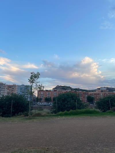 The image depicts an outdoor scene with a view of buildings and a cloudy sky. In the foreground, there is a dirt or gravel area. Behind this, there is a line of trees and vegetation, followed by a chain-link fence. Beyond the fence, apartment buildings of various colors, including orange, blue, and grey, are visible. Further in the distance, on elevated terrain, there are structures that appear to be ancient ruins or fortifications. The sky is a mix of blue and white clouds, with some areas illuminated by sunlight, suggesting it might be late afternoon or early evening. The overall impression is of a cityscape viewed from a park or elevated open space, possibly in Sagunto, Spain, given the mention of fortifications on the hills which are characteristic of the Roman city's historical sites. There are no people clearly visible in the image.