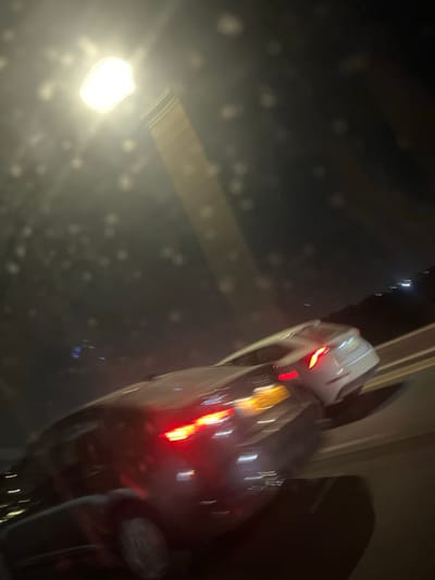 The image displays a blurry, nighttime street scene in Lod, Israel, photographed through a medium exhibiting numerous light refractions, consistent with a wet or dirty surface. A prominent, bright white light source is positioned in the upper-left quadrant, with a linear light-colored structure extending downwards from it. Two vehicles are visible on a multi-lane roadway, both exhibiting motion blur. A dark-colored vehicle occupies the immediate middle ground, displaying illuminated red tail lights and blurred amber light streaks originating from its rear. Behind this vehicle and slightly to the right, a lighter-colored vehicle is partially visible, also showing illuminated red tail lights. The cars appear to be traveling in the same direction. The foreground includes dark shapes consistent with the interior elements and a wheel of the photographing vehicle. No individuals are discernible within the frame.