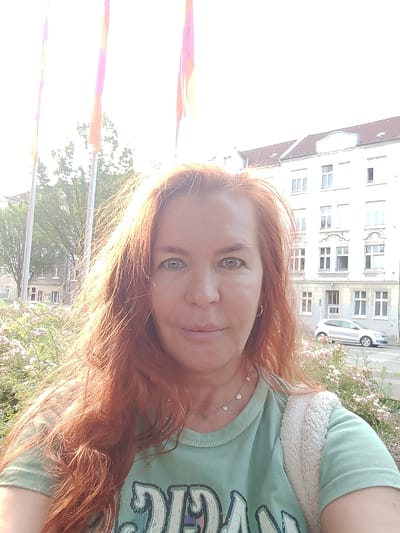 A woman with long, red hair is taking a selfie outdoors, with a building and trees in the background. She is wearing a light green t-shirt and a necklace. A white fluffy bag is slung over her shoulder. The lighting suggests it is daytime, possibly late afternoon due to the warm glow on her hair. The building in the background appears to be an apartment building with multiple windows. A silver car is parked on the street in front of the building. There are also some flowering bushes in the foreground. The overall scene suggests a casual outdoor setting. It is not possible to determine the specific location in Dortmund, Germany from the visual information provided.