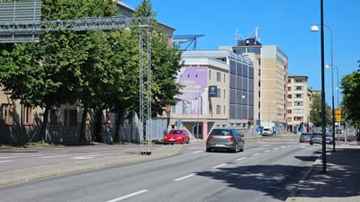 The image captures a street scene in Linköping, Sweden, with several cars driving on a paved road. The buildings lining the street are a mix of modern and older architectural styles, with apartment buildings and commercial spaces. Trees with lush green foliage are present along the sidewalks and between some of the buildings. The sky is clear and blue, indicating a sunny day. A large billboard with a purple background and advertising for "dukatten" and "LinPark" is visible on the side of a building. Traffic lights and streetlights are also present. One car, a red Volkswagen Golf, is stopped at a traffic light, while other cars are moving or parked. The sidewalks are paved with cobblestones and there are shadows cast by the trees and buildings, indicating the time of day is likely afternoon.