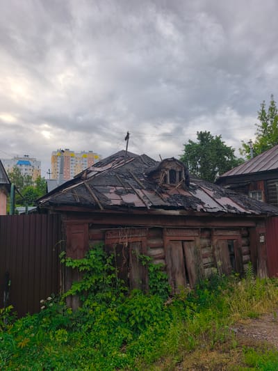 This media file depicts a close-up, eye-level view of a dilapidated wooden house with a damaged roof, set against a backdrop of modern apartment buildings. The house, which appears to be abandoned, is made of logs and has weathered wooden siding. The roof is severely damaged, with large sections missing and exposed wooden beams. A dormer window on the roof is also in disrepair. Lush green vegetation, including vines, is growing up the side of the house, further emphasizing its neglect.
To the left of the house, a dark brown metal fence runs along the property line. In the background, several multi-story apartment buildings are visible, some with colorful facades and balconies, indicating a contrast between the old and the new in this location. The sky is overcast with heavy, gray clouds, suggesting a cloudy or possibly rainy day. The lighting is diffused, and there is no direct sunlight. The scene evokes a sense of decay and abandonment, juxtaposed with the signs of modern life in the background.
There are no people visible in the image. The activity taking place is passive; the image captures a static scene of a decaying building in a residential area.
Notable details include the visible number "68" on the side of the house, possibly a house number. The time of day is difficult to determine precisely due to the overcast sky, but it appears to be daytime. The location is identified as Tula, Russia, which provides geographical context. The overall mood conveyed is one of abandonment and the passage of time.