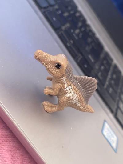 A toy Spinosaurus dinosaur rests on a closed laptop. The laptop sits on a pink surface, potentially fabric. The background shows a blurred laptop keyboard and a label affixed to the laptop's surface. There is no indication of Elena, Bulgaria in the image.