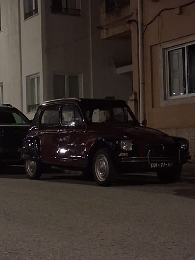 A dark maroon classic automobile with license plate "DM-71-50" is parked on the street, partially obscured by another dark vehicle. The background shows the facade of a building with several windows. It is nighttime, and the lighting appears to be ambient street lighting. The location is Figueira da Foz, Portugal, based on the license plate code.