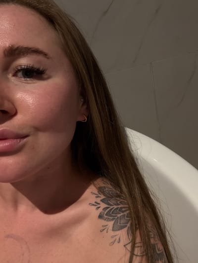 A close-up, angled photograph depicts a woman's face, neck, and upper chest. She has light skin, brown hair, and a tattoo of a geometric floral design on her shoulder. A gold earring is visible in her ear. Part of a white, curved object, possibly a bathtub, is visible in the background. The background also includes a textured, light-colored wall. The setting is a bathroom, but there is no indication of Zhangjiajie, China.