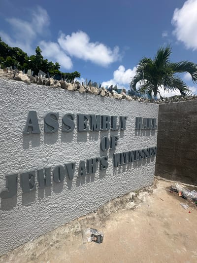 The image shows a textured white wall with raised grey letters spelling "ASSEMBLY HALL OF JEHOVAH'S WITNESSES". The wall is topped with broken glass embedded in concrete, a common anti-climbing security measure. To the left, lush green foliage is visible, and to the right, a palm tree sways against a blue sky with scattered white clouds. The ground in front of the wall is a dirt and concrete surface with scattered debris, including plastic bags and wrappers. The image is taken from a low angle, looking up towards the wall and sky. The context provided by the user identifies the location as Itoga, Nigeria.