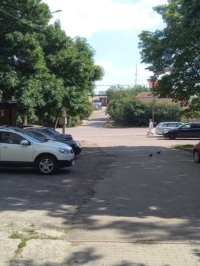 A street scene in Rivne, Ukraine, features parked cars and a pedestrian. In the foreground, a white SUV is parked on a gravel lot, to its right is a black sedan, also parked. Beyond these vehicles, a paved intersection is visible. A person, appearing to be a woman with reddish hair, walks across the intersection carrying a light-colored bag. Several trees with dense green foliage line the street. The sky is a clear blue with a few wispy clouds. The overall impression is of a typical summer day in an urban environment.