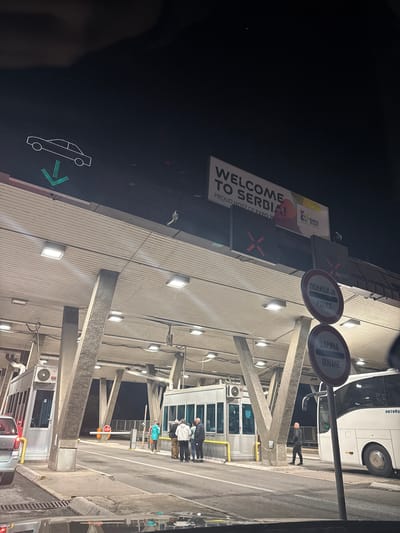 The image captures a nighttime scene at a modern border crossing facility, likely in Dimitrovgrad Municipality, Serbia, illuminated by numerous square-shaped lights mounted on the concrete structure. The sky is dark, emphasizing the artificial lighting.

On the left, a large billboard proclaims "WELCOME TO SERBIA! PROUD HOST OF EXPO 2027 BELGRADE SERBIA." Below this, a digital sign displays a red 'X', indicating a closed lane, while further left, another digital sign shows a white car symbol and a green arrow pointing right, signifying an open lane for vehicles.

The central part of the image features robust concrete pillars supporting the overhead structure, which houses multiple processing booths. Two prominent circular signs are visible: one reads "ПОЛИЦИЈА POLICE" and the other "ЦАРИНА DOUANE" (Customs), indicating the functions of the checkpoint. Several people are present, appearing to be travelers or officials. One person stands next to a large white bus, partially visible on the right, which has "автобус" (bus) written on its side. A group of three individuals is seen standing near one of the booths further into the facility, and another person is near a booth in the foreground.

Barriers are present across the lanes, with one displaying a red "STOP" sign. The ground is asphalt, showing lane markings. The overall setting is a functional and brightly lit border control point, with activities related to vehicle and passenger processing taking place.