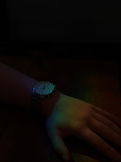 A close-up shot captures a left human forearm and hand, resting on a dark wooden surface. A silver wristwatch with a white face is worn on the wrist. A band of colorful light, resembling a rainbow or prism effect, illuminates the forearm and hand. The background is predominantly dark and out of focus, creating a stark contrast with the illuminated subject. There is no discernible location within Ipswich, United Kingdom, as the scene is an indoor, undetailed environment.
