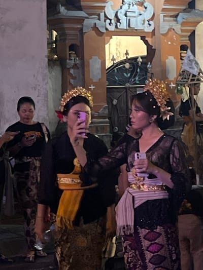 The scene depicts an outdoor gathering at night in Munggu, Indonesia, set against the backdrop of an ornate traditional Balinese building facade with orange/peach walls and lighter decorative carvings, featuring a dark metal gate partially visible in the background.
In the foreground, two young adult females are prominent, both wearing elaborate golden floral headpieces and traditional Balinese attire. The woman on the left, facing forward, wears a black long-sleeved top and a patterned sarong with a golden sash; she holds a purple smartphone vertically in front of her face, appearing to record or photograph. A red flower is visible behind her left ear. The woman on the right wears a black lace long-sleeved top with a V-neck, a patterned sarong featuring maroon and pink geometric designs, and a light-colored sash with a striped waistcloth; she holds a white smartphone in her right hand, pointed generally forward. Her gaze is directed slightly to her left.
Behind the two foreground figures, to the left, a middle-aged female with dark hair pulled back wears a black t-shirt with an orange graphic and a patterned sarong. She holds a dark smartphone and looks down at it. Further in the background, to the right, a person is partially visible carrying a tall, conical structure made of a light-colored material, possibly bamboo, adorned with white cloth or paper. Another partial figure is visible on the far left edge. The scene is illuminated by artificial light, casting warm tones on the architecture.
