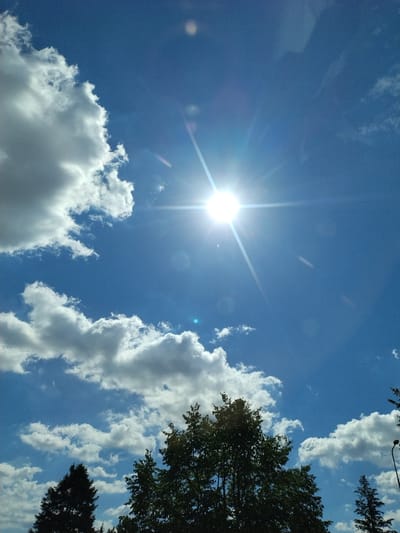 The image captures a clear, bright blue sky with scattered white cumulus clouds. The sun is prominently visible, slightly off-center, casting a bright glare and visible sunbeams. In the lower portion of the image, the dark silhouettes of trees are visible against the sky. The overall scene suggests a sunny day. Without further context or identifying markers, it is not possible to determine the specific location within Strakonice, Czechia.