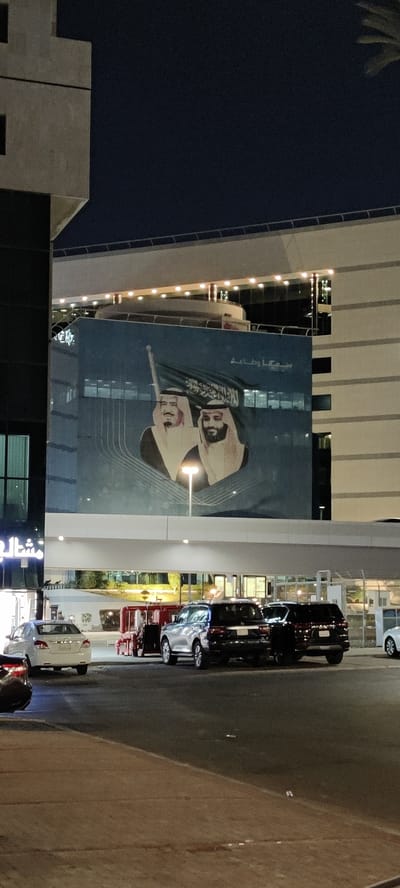 This night-time photograph captures a vibrant urban scene in Jeddah, Saudi Arabia. A large, modern building dominates the background, its blue-tinted facade featuring a prominent banner. This banner displays portraits of two distinguished figures, identified as King Salman bin Abdulaziz Al Saud and Crown Prince Mohammed bin Salman Al Saud, set against the green backdrop of the Saudi Arabian flag. Above the portraits, Arabic text reads "سمعاً وطاعة" (Sami'an wa Ta'ah), translating to "Listen and Obey" or "Hearing and Obedience," a common expression of loyalty.
The street level in front of the building is active with several parked cars, including a white sedan, a silver SUV, and a dark-colored SUV. Bright artificial lighting illuminates the scene, with a prominent light source directly below the banner, enhancing its visibility. Building interiors and other streetlights also cast light on the surroundings. A paved street and a concrete sidewalk are visible in the foreground. On the left side of the building, partially visible Arabic text, possibly "مشالح" (Mashalih), suggests a commercial establishment. The clear night sky indicates favorable weather conditions, making for a well-lit and discernible urban landscape. The overall impression conveys a sense of national pride and a typical bustling night in the city.