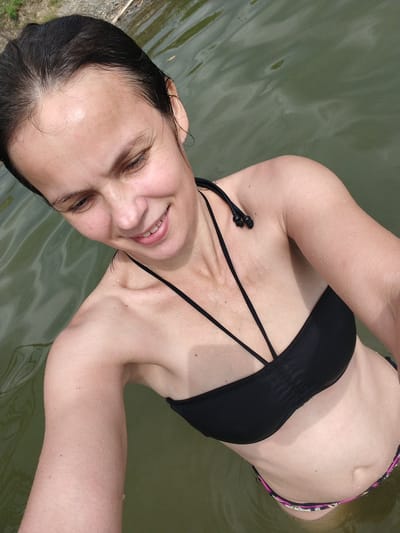 A young woman with dark hair, wet from swimming, smiles at the camera as she stands in a body of water. She is wearing a black bikini top with thin straps and a colorful bikini bottom. Water droplets are visible on her skin. The water appears murky green with ripples on its surface, suggesting it is a natural body of water like a lake or river. The background shows a blurry view of what appears to be foliage and a log, indicating a natural outdoor setting. The lighting suggests it is daytime, likely a warm day, given the woman is swimming. She appears happy and relaxed. There is no visible text in the image.