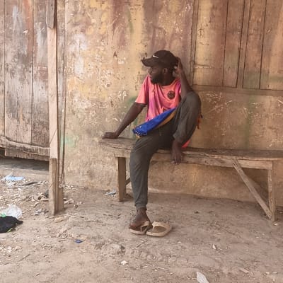 An adult male is seated on a weathered wooden bench in Kaura Goje, Nigeria. He is positioned against a distressed, light brown wall featuring peeling paint and a visible patch of green discoloration. To the left, a vertically paneled wooden door or shutter is partially visible. The ground consists of dry, uneven dirt littered with various pieces of trash, including plastic fragments and a white plastic bag. The male, with dark skin and a beard, wears a black baseball cap with a white logo, a pink short-sleeved t-shirt with a club crest (likely Paris Saint-Germain), and dark-colored track pants. A blue and yellow striped fabric item is draped over his left thigh. His right hand is raised to his head, appearing to adjust his cap or hair, while his left arm rests on the bench to his side. His right leg is bent with the foot on the bench, and his left leg extends towards the ground. A pair of light brown flip-flops are present, one partially on his left foot, the other resting on the ground nearby. He is looking towards the left of the frame. The scene is illuminated by direct daylight.