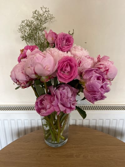 A bouquet of pink and purple peonies and roses sits in a clear glass vase on a wooden table. The vase is filled with water, and the stems of the flowers are visible. In the background, a white radiator is visible, and behind that, a textured off-white wall. The lighting is soft and natural. The image appears to be taken indoors, possibly in a home or a shop. There is no indication of the location being in Portarlington, Ireland.