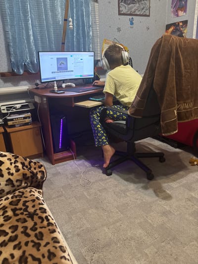 This is a vertical, high-angle shot of a young person sitting at a desk in what appears to be a bedroom. The person is wearing a yellow t-shirt, blue and yellow patterned pajama pants, and white headphones. They are looking down at a computer monitor, suggesting they are engaged in an activity such as gaming or browsing the internet. The computer tower under the desk has a purple light emanating from it.
The desk is made of wood and has a monitor, keyboard, mouse, and a white phone. On the wall behind the desk, there are several posters or framed pictures. A brown, fuzzy blanket is draped over a red chair next to the desk. A portion of a leopard-print sofa is visible in the foreground on the right. The room is carpeted with a light gray, patterned rug. Blue curtains are visible to the left of the desk, with a fluorescent light fixture above them.
No specific time of day or weather conditions can be determined. The setting appears to be a residential room. Some visible text on the monitor shows a website with options related to downloading images. There are also books on a shelf below the desk, one of which has Cyrillic text, likely indicating the location as Ukraine.