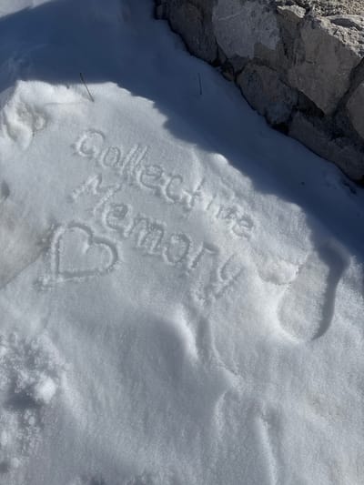 The image depicts a snow-covered ground surface, adjacent to a dry-stacked stone wall, located in Virak, Montenegro. Inscribed into the snow, through shallow depressions, are the words "Collective Memory" arranged in two lines. Below this text, a heart shape is also drawn into the snow. Two thin, dark-colored sticks protrude from the snow: one near the upper left quadrant and another closer to the stone wall on the right. The stone wall is constructed from irregularly shaped, light-grey to dark-grey stones. Direct sunlight illuminates the scene from the left, casting distinct shadows, including those that define the inscribed text and a diagonal shadow line across the snow. No individuals or active interactions are present in the frame.
