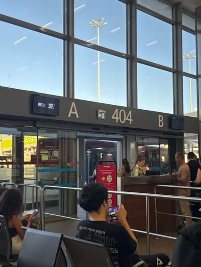 The image depicts an airport gate area, likely in Pendik, Turkey, given the text visible. Several people are waiting at a gate, suggesting an airport or train station. The scene is indoors, with a large window showing a bright, clear sky, indicating daytime and good weather.
The dominant feature of the image is a large, dark gray signpost with white lettering indicating "A", "B", and "404". Below the "A" is a small black screen displaying a logo and text, which appears to be for Istanbul Sabiha Gökçen International Airport. Reflecting on the glass, we can see signs with the word "GOKCEN" and "PEGASUS", further confirming the location.
The people in the image are either standing or seated, mostly looking at their phones. One person in the foreground, wearing a black t-shirt with text, is looking down at their phone. Another person, with curly hair, is also engrossed in their phone. Further back, a woman with red hair is looking towards the gate area. The overall mood seems to be one of patient waiting, typical of travel hubs. The presence of metal railings suggests a queuing system for boarding. The lighting suggests it is daytime, likely sunny.