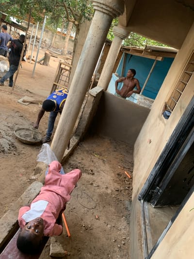 The image depicts an outdoor construction or renovation site in Dekpor Horme, Ghana, under clear daylight conditions. In the immediate foreground, a young individual, possibly a child, wearing a pink patterned tunic with a white chest panel, lies supine on a wooden plank, looking directly at the camera. To the mid-left, a male individual wearing a blue and yellow basketball jersey with "CURRY" and number "30" on the back is bent over, handling materials in a grey circular basin on the dirt ground, indicative of mixing mortar or cement. Further in the background, two additional males are visible; one in a dark shirt and jeans is walking towards the left.

The scene is framed by various architectural elements. On the right, a light tan-colored building wall features a dark-framed opening, possibly a window or doorway. Prominently in the center, a robust concrete column with a capital extends vertically. Another similar column is partially visible to its right. Above the ground level and to the mid-right, a shirtless male with dark skin stands behind a newly plastered, smooth, light grey wall section. He has his hands on his hips and is looking towards the left. Behind him, a structure with light blue-painted walls is visible.

The ground consists of disturbed earth and dirt, with construction debris including small orange cylindrical objects, wood planks, and scattered rubble. A clear plastic bag rests on the ground near the central column. Overhead and in the distant background, green foliage and trees are visible, suggesting a natural outdoor environment surrounding the construction area. The overall impression is one of active building work in a populated, semi-rural or peri-urban setting.