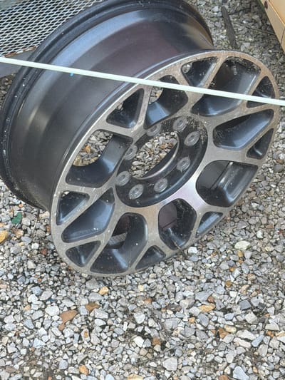 An aluminum alloy wheel, likely from a vehicle, rests on a gravel surface. The wheel is dark gray with silver accents, featuring a complex spoke design with circular cutouts. A light-colored flexible tube is draped across the face of the wheel. The gravel is composed of various small rocks and some dried vegetation, suggesting an outdoor environment. The scene is located in Hornersville, United States.