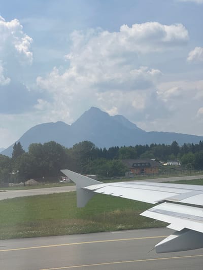 The image shows an airplane wing in the foreground, positioned over a tarmac and grassy area. In the background, a range of hazy mountains dominates the horizon, with a dense forest separating them from a few buildings, including an orange-colored house. The sky is partly cloudy with patches of blue visible. This scene is consistent with an airport environment in or near Salzburg, Austria, given the proximity to the Alps. There are no people visible in the frame. The perspective suggests the airplane is either on the ground at the airport or has just taken off.
