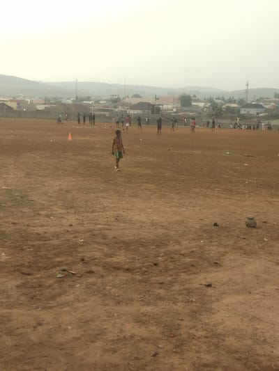 The image shows an open, dirt field in New Karu, Nigeria, at the coordinates 8.979 latitude and 7.565 longitude. The field appears to be used for playing soccer as there are several people playing and there is a small orange cone in the foreground. There is a single boy walking towards the camera in the center of the field. Several other people are playing soccer in the distance, and there is a low-lying hill in the background. The scene is overcast and a small amount of greenery can be seen in the field, along with some trash. There are several houses and buildings in the distance, indicating a residential area.