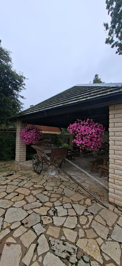 The image depicts an outdoor scene in Kaali, Estonia, featuring a rustic covered seating area. In the foreground, a stone-paved courtyard is visible, with large, irregularly shaped stones forming the ground. To the left, a weathered wooden cart with spoked wheels is prominently displayed. Hanging baskets overflowing with vibrant pink flowers are suspended from the structure. The covered area itself has a dark, sloped roof with visible moss or algae growth. Light-colored brickwork forms a pillar on the right side of the structure, and the background shows glimpses of trees and more stone walls. The overall atmosphere is tranquil and suggestive of a rural or historical setting, likely part of a park or heritage site in Kaali. No people are present in the image.