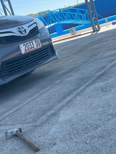 The image shows a partially visible gray Toyota vehicle parked on a light gray concrete surface in Дербісек ауылдық округі, Kazakhstan. The front of the car, including the Toyota emblem and a license plate reading "2033 DV," is in focus. To the right of the car, a trowel lies on the concrete. In the background, a blue metal structure is visible. The scene appears to be outdoors under daylight.