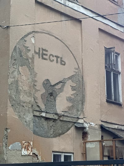 The image shows the exterior wall of a building in Ivano-Frankivsk, Ukraine. A large, circular mural depicting a silhouette of a person holding a rifle is painted on the wall. The word "ЧЕСТЬ" (Honor in Russian) is written in large Cyrillic letters above the silhouette within the circle. The mural is weathered and partially faded, with sections of the plaster peeling away, revealing the underlying brickwork in places. A window with multiple panes and a partially open shutter is visible to the right of the mural. Below the mural, a circular sign with the number "4/3" is attached to the wall. The building appears to be aged, with visible signs of wear and tear on its facade. The sky is overcast, suggesting a cloudy day.