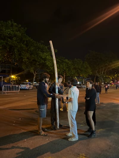Four individuals are gathered outdoors at night, near a street with trees lining the background. The main focus is on a tall, cylindrical object, possibly a pole or a large pipe, held vertically by two men in the foreground. One man, with dark hair and a beard, wears a dark long-sleeved shirt and shorts, and sandals. He is shaking hands with another man, who is wearing a black leather jacket over a lighter shirt and dark pants. To the right, a third person in a striped shirt and light-colored pants is also present, facing the two men. A fourth individual, a woman in a black dress and boots, stands to the far right, looking towards the group. In the background, a police car with illuminated blue and white markings is visible, as well as metal barriers and more people in the distance. The scene appears to be in a public space, possibly a street or plaza, with streetlights casting warm illumination and creating long shadows on the asphalt. The location is identified as Matosinhos, Portugal.