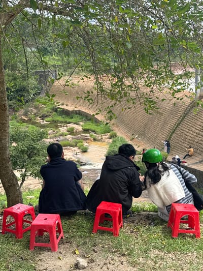 The image depicts a scenic overlook in Lang Biang - Da Lat, Vietnam, featuring a group of people enjoying the view of a rocky riverbed and a large, terraced embankment. In the foreground, three individuals are seated on the grass, facing away from the camera. The person on the left wears a dark blue jacket, the middle person a black jacket, and the person on the right is wearing a white and gray striped sweater and a green helmet. In front of them are three bright red plastic stools. The background reveals a watercourse with flowing water and exposed rocks, surrounded by lush green vegetation. Further in the distance, the terraced embankment dominates the landscape, with several figures visible walking and standing on it. The scene is framed by the branches of a large tree in the upper portion of the image.