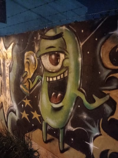 The image shows a graffiti mural on a wall in El Talar, Argentina. The mural depicts a green, humanoid character with one large eye, a wide open mouth, and an outstretched arm holding a yellow, circular object. The character appears to be laughing. The background is a dark, starry sky. The mural is located on a beige wall with green plants growing at the bottom. There are no other people or objects visible in the image. The mural is located at latitude -34.462 and longitude -58.652.