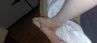 A foot is being held by a hand. The foot has purple nail polish on the big toe. The hand and foot are on white sheets and a tan blanket. A purple wristband is on the wrist. A wooden floor is visible. A black metal stand is visible in the background. It is dark in the room. It is not possible to determine the location in Fort Smith, United States of America from the image.