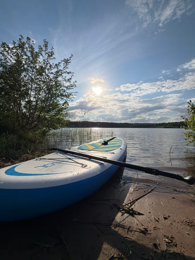 The image shows a paddleboard lying on a sandy shoreline next to a lake or river. The paddle is resting on the board. The sky is visible, with clouds and sunshine. Trees and vegetation are visible along the shoreline. The text on the paddleboard reads "105 SOL". It's impossible to determine the location is Selychka, Russia from the image alone.