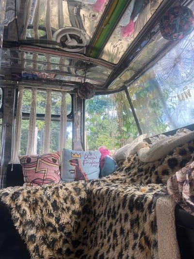 The image shows the interior of a vehicle, possibly a bus or jeepney, in Thong Sala, Thailand. The interior is adorned with patterned pillows, a leopard-print blanket, and reflective metal trim. The windshield is cracked. Outside, there's green foliage and trees.