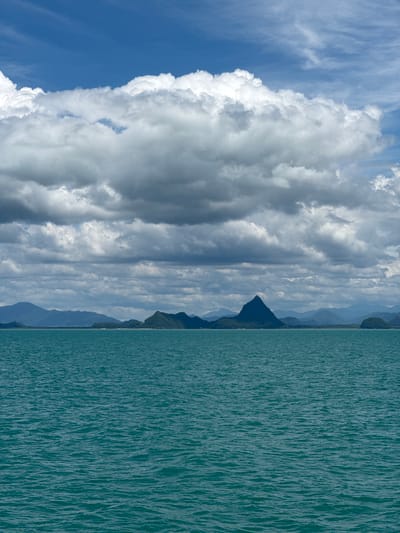 This is a vertical, eye-level shot of a body of water meeting a distant island chain under a cloudy sky. The foreground is filled with the deep teal-colored ocean, its surface rippled by gentle waves. In the middle ground, a long chain of green, mountainous islands stretches across the horizon, with a prominent, sharply peaked island in the center. The background consists of a layered sky with dense, dark gray clouds filling the lower portion and lighter, fluffier white clouds in the upper portion, broken by patches of bright blue sky. The scene is devoid of any people or man-made structures. The overall impression is one of natural, serene, and slightly dramatic coastal scenery.