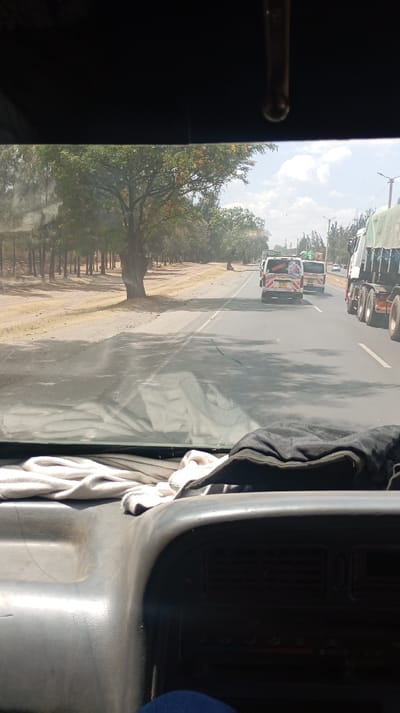 The view from inside a vehicle shows a road with trees lining one side. Several vehicles, including vans with distinctive markings and a large truck, occupy the road ahead. The vehicle's dashboard is visible in the foreground, cluttered with a white cloth and dark material. Sunlight streams through the windshield, creating shadows on the road.