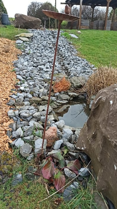The scene depicts an outdoor landscaped area in Florenville, Belgium, under an overcast sky. A linear bed, filled with irregularly shaped grey rocks and small stones, extends diagonally from the lower left towards the upper right of the image. To the immediate left of this rocky bed, an area is covered with light brown wood chips. In the foreground, spanning the bottom edge, are patches of green grass interspersed with dark green and reddish-tinged leaves of low-growing plants.
Centered within the rocky bed in the foreground stands a tall, slender, rust-colored metal stake. It is topped with a shallow, bowl-shaped receptacle, on which rests a small, dark, bird-shaped figurine. Near the base of this stake, among the rocks, lies a smaller, reddish-brown, rounded figurine, possibly depicting a frog or toad.
To the right of the rocky bed, a small, dark water feature or pond is visible, its surface reflecting the sky. This pond is bordered on its right by a large, irregularly shaped, dark brown boulder and backed by an area of dried, light brown plant stalks.
In the mid-background on the left, a large, dark brown boulder and a dark grey cylindrical container (possibly a compost bin) are situated on a grassy slope. In the mid-background on the right, a wooden pergola structure with a dark roof and exposed timber framework is visible, positioned over a manicured green lawn. No human or animal subjects are present.