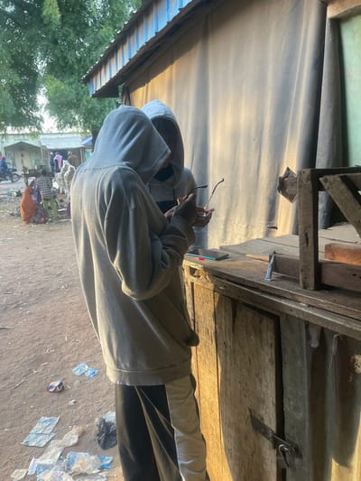 A person in a grey hoodie stands outside under a tin roof, holding glasses. Debris litters the ground, including discarded plastic. In the background, people gather, some near a building. Trees are visible beyond. An old wooden structure with a door is partially visible overhead.