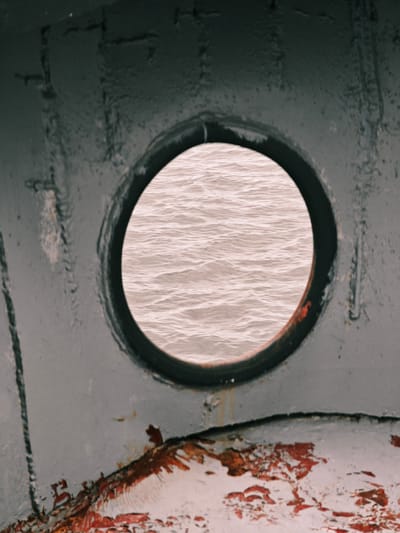 A close-up, weathered view through a porthole of a ship reveals choppy, pale pinkish-grey water. The porthole itself is circular and set within a dark, textured metal frame. The outer hull of the ship is visible in the foreground, painted a dull, dark grey with patches of rust and peeling paint, particularly along the bottom edge. The texture of the metal suggests age and exposure to the elements. The overall impression is one of a utilitarian, possibly aged vessel against a muted aquatic backdrop. The image's composition is tight, focusing on the contrast between the enclosed view through the porthole and the rough, decaying surface of the ship. No people or specific location in Zotino, Russia, are discernible from this image.
