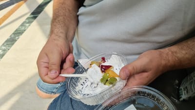 The image shows a person holding a clear plastic container of whipped cream and fruit salad. The person is wearing a light blue polo shirt and blue jeans. The container is partially filled with whipped cream and topped with pieces of fruit, including kiwi, what appears to be peach or apricot, and possibly pomegranate seeds or dried cranberries. A white plastic spoon is inserted into the whipped cream, and a piece of whipped cream and fruit has been scooped onto it.
The background shows a floor with a pattern of cream-colored and colored stripes, possibly marble tiles. The lighting suggests it is daytime. The setting appears to be indoors or in a shaded outdoor area, given the even lighting and absence of harsh shadows. The overall impression is casual, possibly a snack break or dessert. There is no visible text that is clear enough to read. The location context is Taranto, Italy.