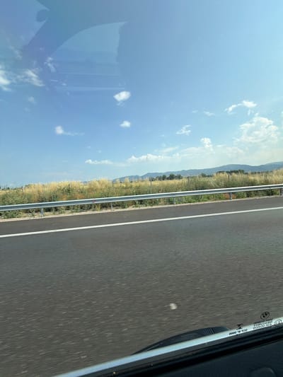 A photograph taken from inside a vehicle shows a highway with dry, golden grass and vegetation lining the side of the road. In the background, rolling hills covered in trees can be seen under a clear blue sky with scattered white clouds. The road surface is dark asphalt with a white line marking the lane. The reflection of the sky and clouds is visible on the windshield of the vehicle. There are no people or animals visible in the image. This scene is consistent with the landscape found along the road near Gorni Bogrov, Bulgaria.