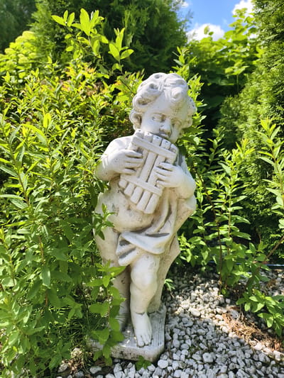 A full shot captures a white cherub statue holding a pan flute in a garden setting. The statue is positioned in the center of the frame, with lush green foliage surrounding it. The cherub is depicted as a plump infant with curly hair, wearing a drapery that covers its lower body. Its hands are clasped around the pan flute, which rests against its chest. The statue stands on a base of small, white gravel. The background consists of dense green bushes and trees, suggesting a well-maintained garden. The lighting indicates a sunny day, with shadows cast by the foliage and the statue. The scene appears to be located outdoors, likely in a residential garden or park area. This image is from Malaya Shil'na, Russia.