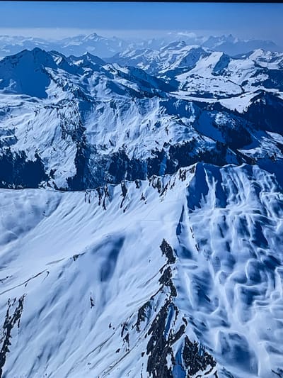 A majestic, wide shot captures a vast expanse of snow-covered mountains under a clear daytime sky. The image is dominated by cool tones of blue and white, showcasing rugged and jagged peaks, steep slopes, and dramatic ridgelines. Atmospheric haze renders distant mountain layers in lighter shades of blue on the left side, enhancing the sense of depth and scale.

The snow cover is extensive, revealing intricate textures and patterns formed by wind and natural processes. Some areas appear smooth, while others are sculpted with wavy drifts and sharp contours. Dark, exposed rock formations, often spiky or cliff-like, punctuate the white landscape, providing stark contrast, especially along the upper ridges and steeper faces.

A notable subject within this grand natural setting is a dark, sleek aircraft, appearing to be a military jet, flying low over the right-hand side of a snow-covered mountain slope. Its silhouette is distinct against the white snow, suggesting it is maneuvering through the challenging mountainous terrain.

The scene depicts clear weather conditions with ample sunlight, indicated by crisp shadows and good visibility across the range. The absence of clouds around the peaks suggests a calm, bright day. The overall impression is one of remote, untouched wilderness intersected by a powerful, modern technological presence. Given the location context, this scene likely takes place in the Carpathian Mountains of Romania, possibly involving an aircraft from the Otopeni region. No text is visible in the image.